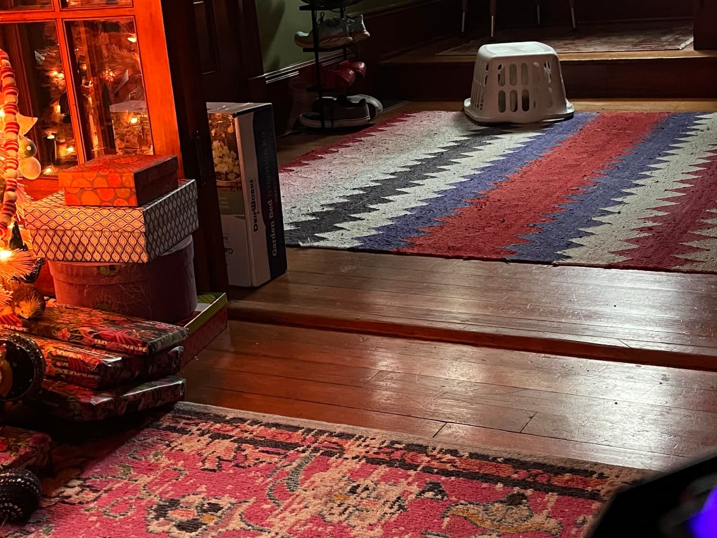 A cozy interior space featuring a wooden floor with area rugs, visible stacks of wrapped gifts beside a small Christmas tree, and a white laundry basket placed on a colorful zigzag-patterned rug.