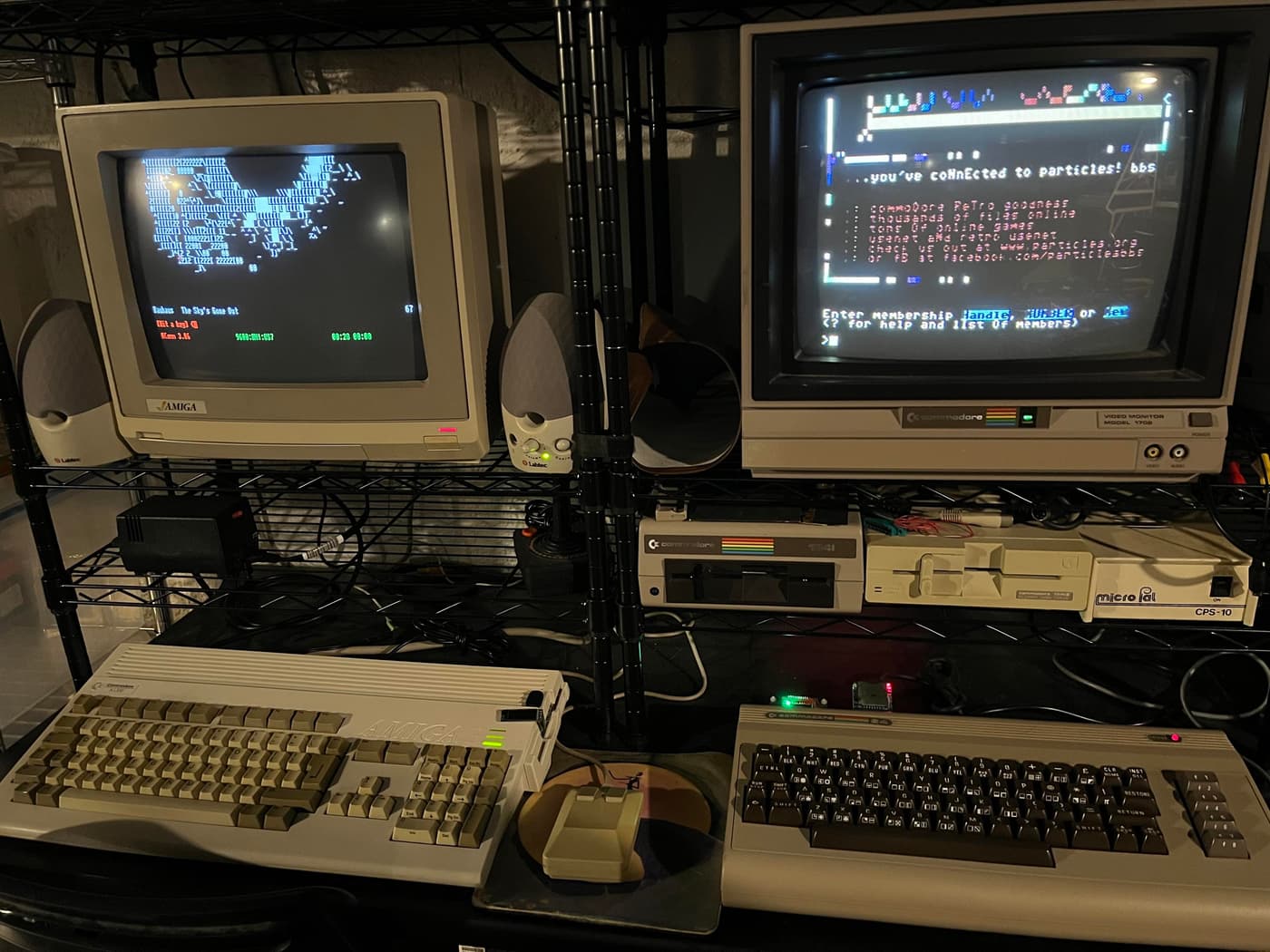 Dual retro computing setup: Amiga 1200 on left showing ASCII skull, C64 on right displaying Particles! BBS with ANSI art, vintage hardware between them