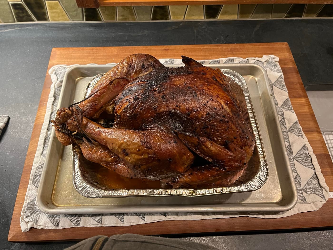 A roasted turkey sits on a silver tray, surrounded by juices, on a wooden cutting board. The turkey has a dark, crispy skin and is positioned with its legs tucked. A patterned cloth is beneath the tray, and the background features a dark countertop