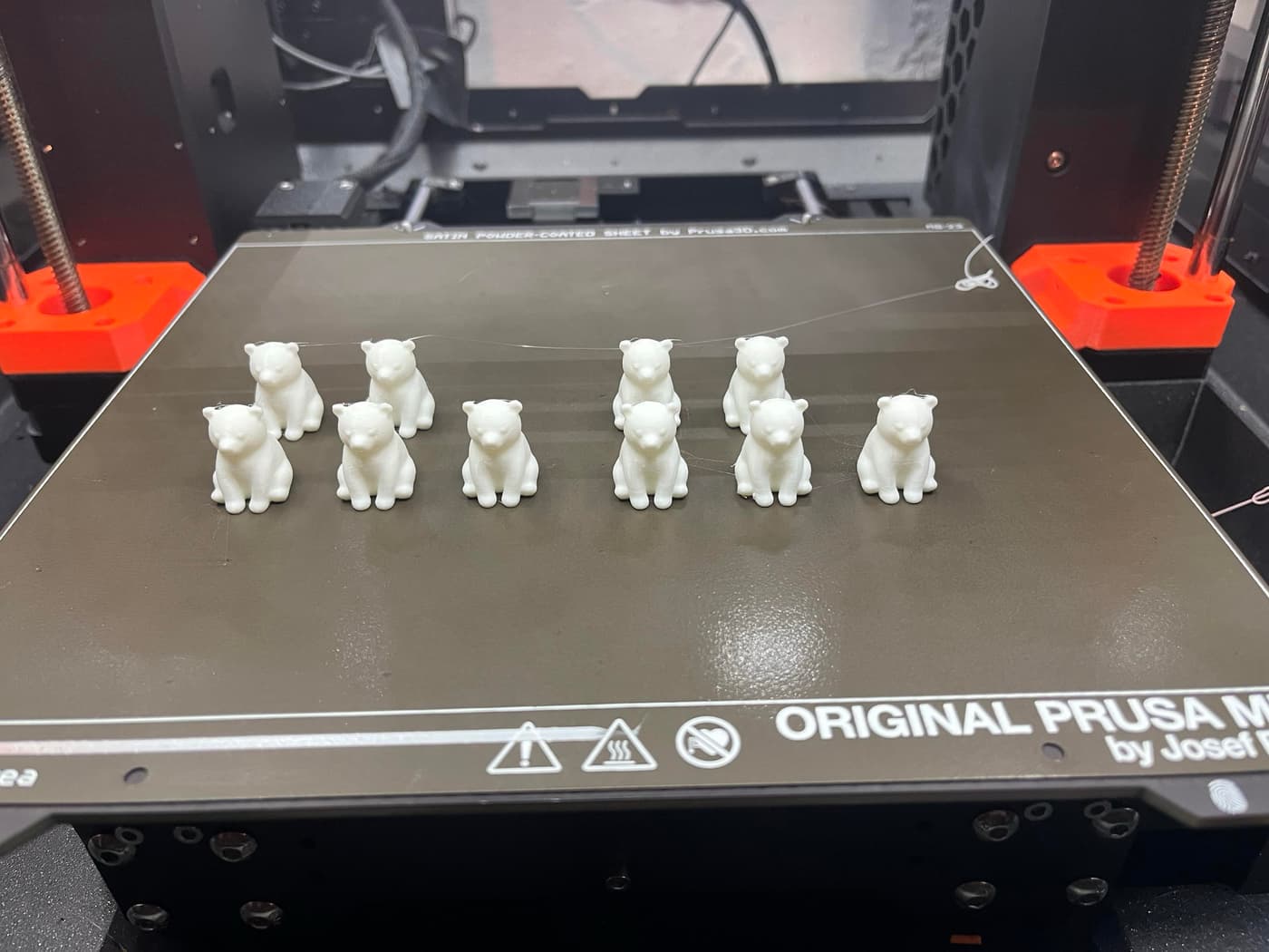 Image features a 3D printer's build plate with ten small white bear figurines arranged in two rows, set against a black background.