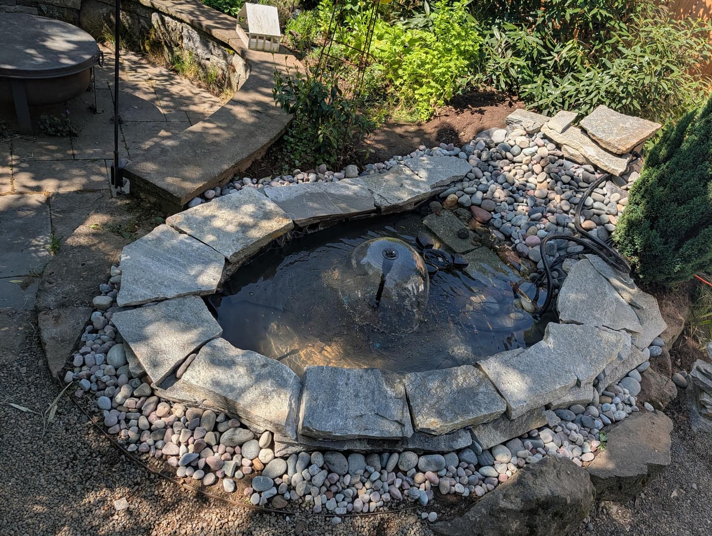 A backyard garden pond with a bubbling fountain, edged with large stone slabs and river rocks