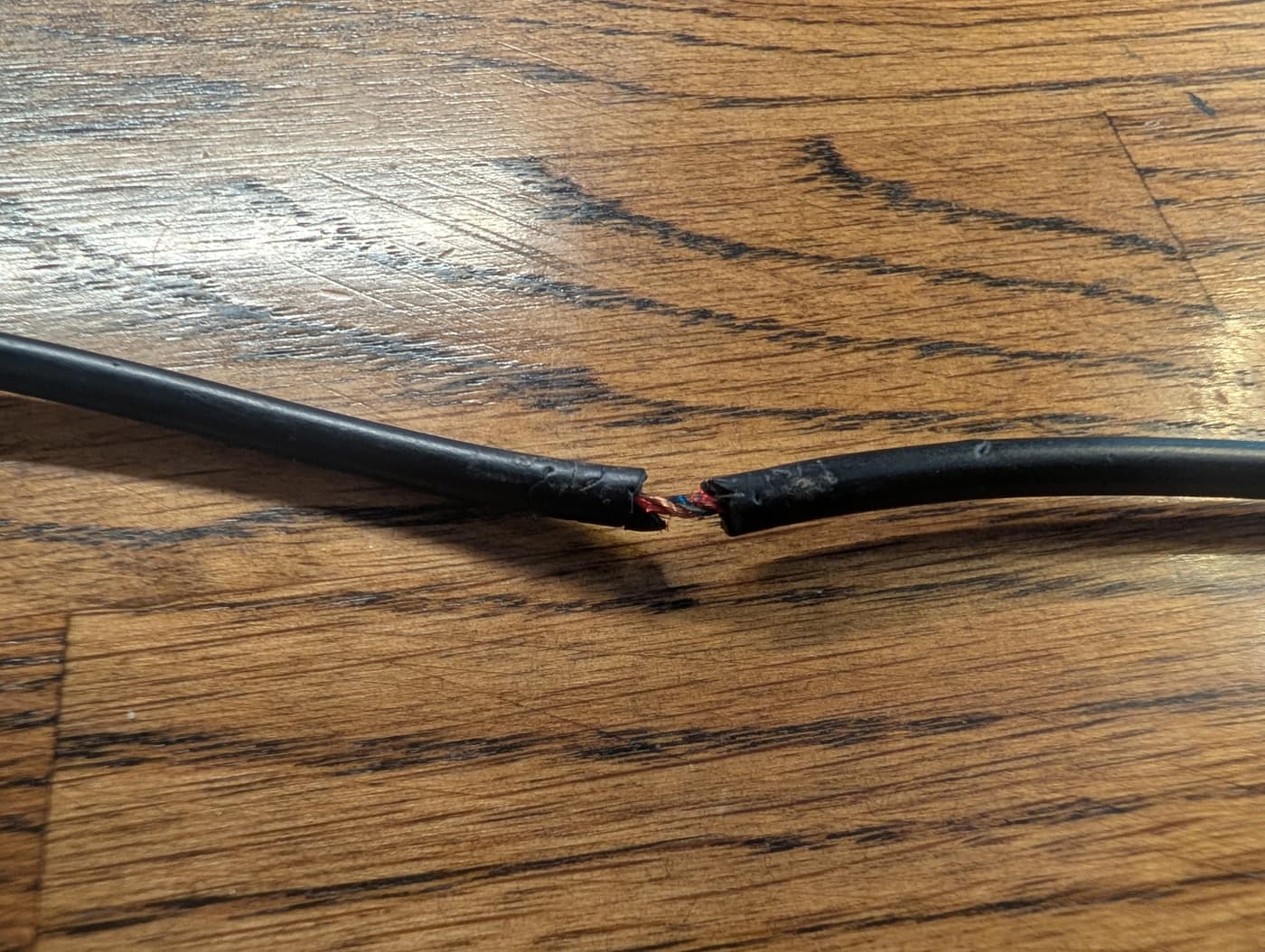 A frayed black electrical cord with exposed copper wires on a wooden surface.