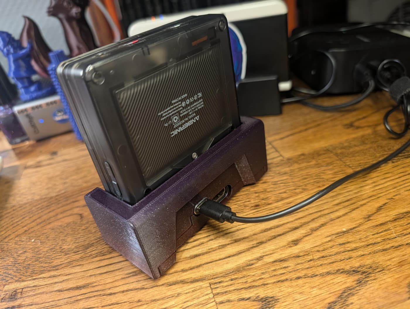 An Anbernic handheld gaming console with a transparent case charging in a purple 3D-printed dock on a wooden desk.