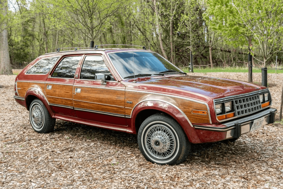 A red 1980s AMC Eagle station wagon with woodgrain side panels and a roof rack, parked on a wooded area.