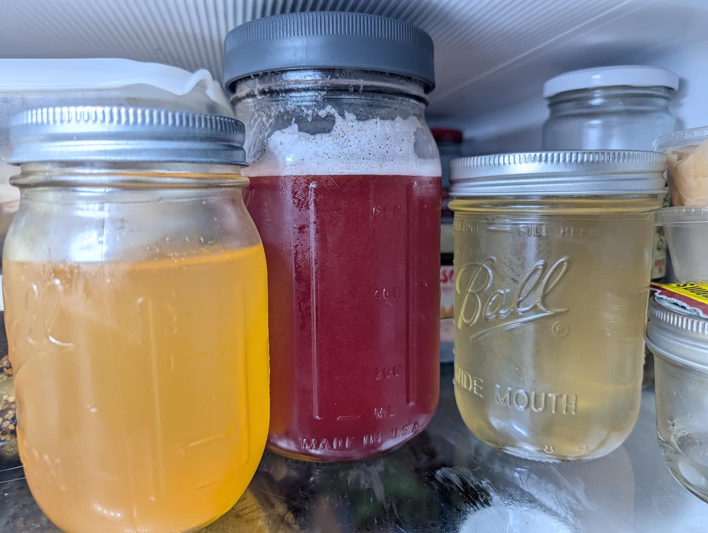 Three mason jars in a refrigerator, including one with yellow liquid and another with red, foamy liquid.